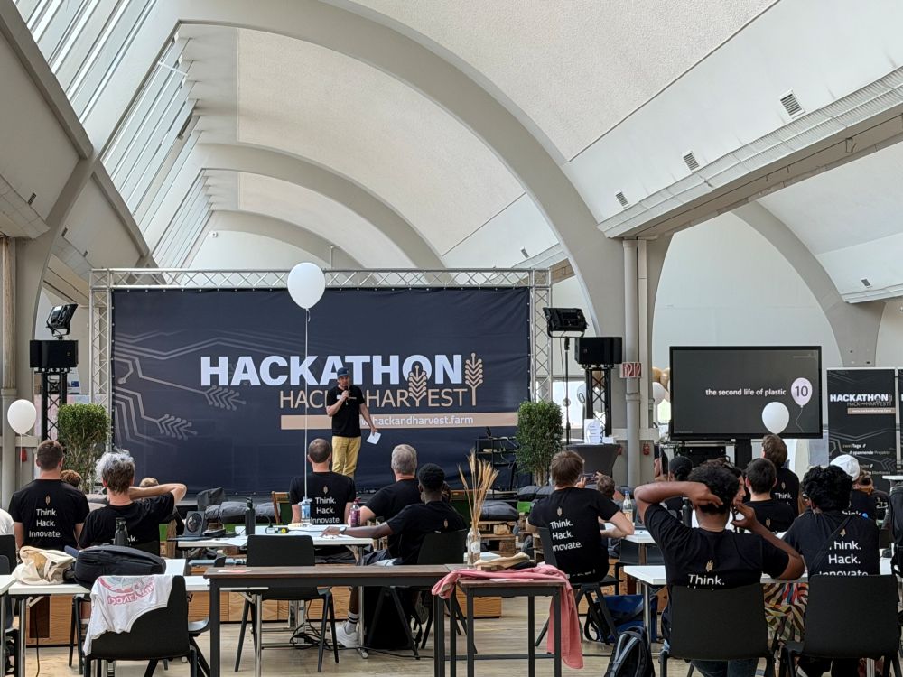 Pitch beim Hack and Harvest im Technologiezentrum Konstanz vor der Bühne mit dem Banner „Hackathon Hack and Harvest“, Teilnehmer in „Think. Hack. Innovate.“-Shirts